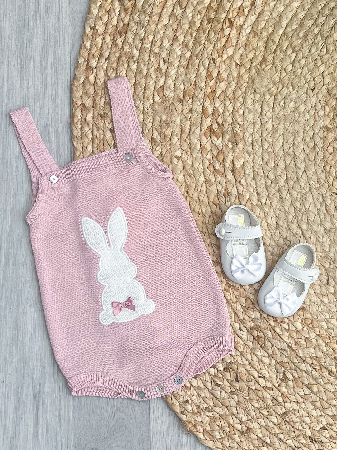Blush pink bunny romper with white bow pram shoes on a brown wicker rug.