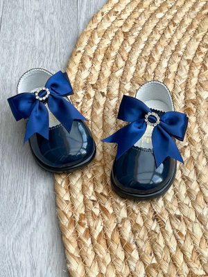 Navy patent leather shoes with a large satin bow to the strap & diamanté ring to the centre on a wicker rug.