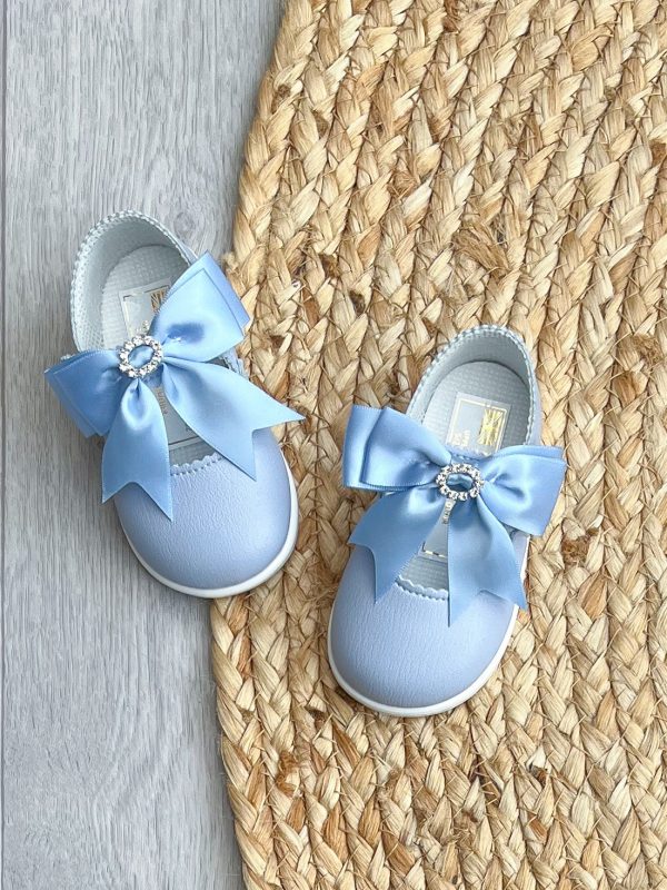 Photo from above of Blue patent leather shoes with a large satin bow to the strap & diamanté ring to the centre on a wicker rug.