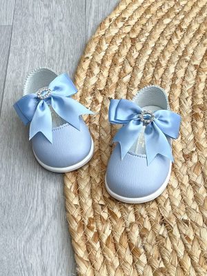 Blue patent leather shoes with a large satin bow to the strap & diamanté ring to the centre on a wicker rug.