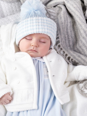 Sleeping baby on a grey blanket, wearing a white knitted jacket & pom pom hat.