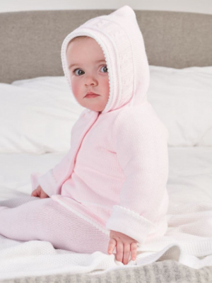 Baby sitting looking at the camera in a pink & white knitted jacket with hood up.